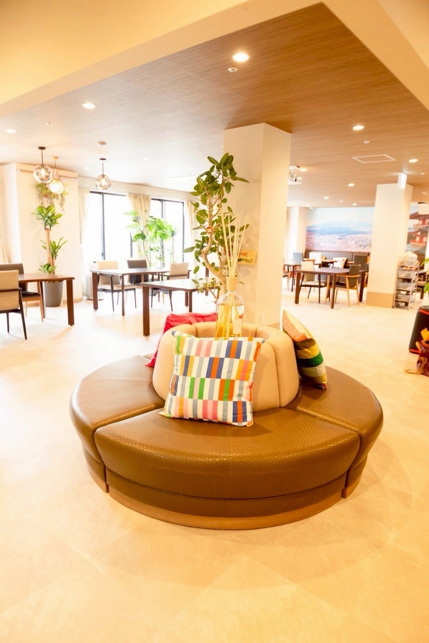 観葉植物に囲まれた円形ソファのラウンジ。夏も涼しい室内で入居者同士が過ごせる(メディケア縁 近鉄八尾駅前)
